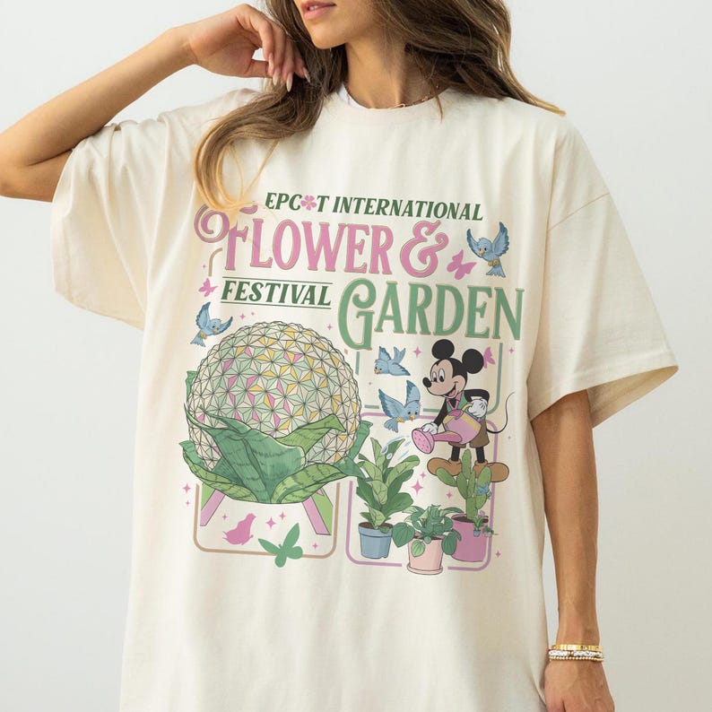May include: Cream-colored t-shirt featuring the text "EPCOT International Flower & Garden Festival." The design includes a geometric sphere, Mickey Mouse, and various plants and butterflies. The shirt has short sleeves and a relaxed fit.