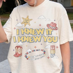 May include: A cream-coloured t-shirt featuring a graphic print with the text "I KNEW IT I KNEW YOU". The design includes a sheriff star with "INFINITY AND BEYOND", a cowboy hat, boots, a beaded bracelet with "You've got a friend in me", and blocks.