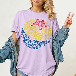May include: A light purple t-shirt featuring a large circular graphic. The graphic is divided into three sections: a red star, a yellow center, and a blue bottom. The design is filled with various small shapes and figures. The shirt has short sleeves and a crew neck.