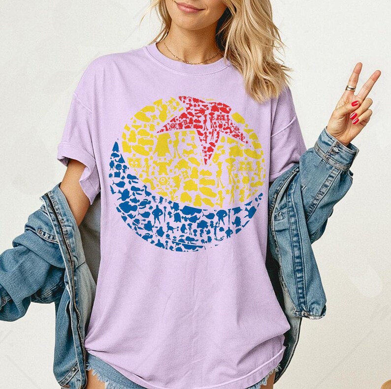 May include: A light purple t-shirt featuring a large circular graphic. The graphic is divided into three sections: a red star, a yellow center, and a blue bottom. The design is filled with various small shapes and figures. The shirt has short sleeves and a crew neck.