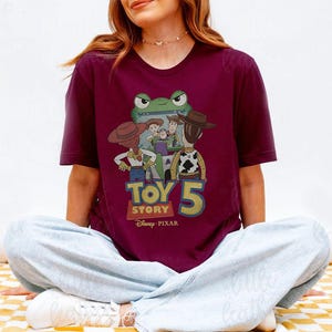 May include: Burgundy t-shirt featuring a graphic of characters from the Toy Story franchise. The design includes the text "TOY STORY 5" and the Disney Pixar logo. The shirt has short sleeves and a crew neck.