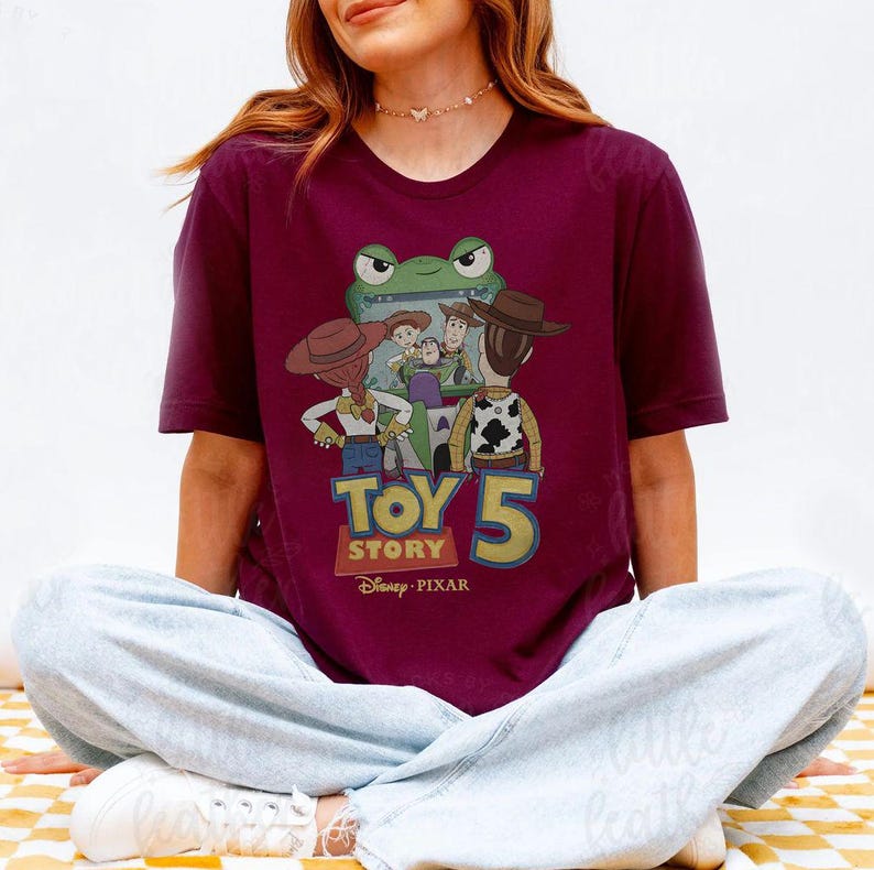 May include: Burgundy t-shirt featuring a graphic of characters from the Toy Story franchise. The design includes the text "TOY STORY 5" and the Disney Pixar logo. The shirt has short sleeves and a crew neck.