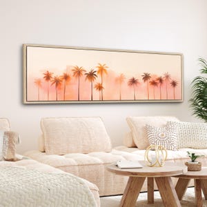 May include: A long, horizontal painting of a sunset scene with palm trees silhouetted against a pink and orange sky. The painting is framed in a light wood frame and is hanging on a white wall above a light-colored couch.