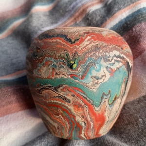 May include: A small, rounded vessel featuring a marbled pattern with swirling layers of red, turquoise, black, and beige. The surface has a matte texture with organic, flowing lines that wrap around the entire exterior of the object.
