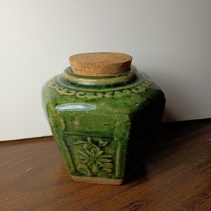 May include: A green ceramic jar with a cork stopper. The jar has a square shape with a floral design on the front and decorative patterns around the top. The jar has a distressed, crackled finish.