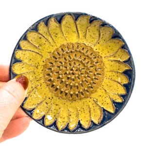 May include: A ceramic dish shaped like a sunflower with a blue rim and a yellow center. The center of the sunflower is textured and appears to be a grater.