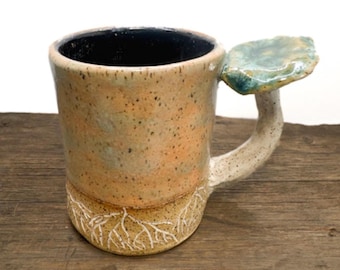 May include: A ceramic mug with a speckled tan and peach glaze, featuring a black interior. The base is decorated with white etched root patterns, and the handle is topped with a flat, textured, green-toned mushroom cap shape.