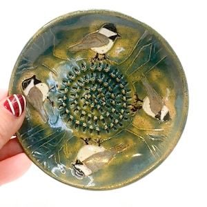 Puede incluir: Un plato de cerámica con un esmalte azul y verde presenta un diseño de cuatro pájaros y un prensa ajos. Los pájaros están pintados en tonos de marrón, blanco y negro.