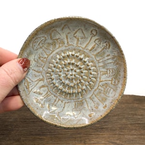 May include: A round ceramic dish with a speckled light gray and tan glaze. The center features a raised spiral pattern of small dots, surrounded by an embossed ring of various mushroom shapes etched into the surface of the clay.