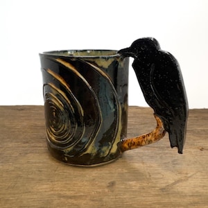 May include: A ceramic mug with a glossy black glaze featuring a carved spiral pattern on the exterior. The handle is shaped like a black bird perched on a textured, light brown branch that extends from the side of the cylindrical vessel.