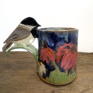 May include: A ceramic mug with a dark blue glaze featuring hand-painted pink coneflower blossoms and green foliage. The handle is uniquely shaped as a curved green branch topped with a sculpted, hand-painted chickadee bird with a black cap.