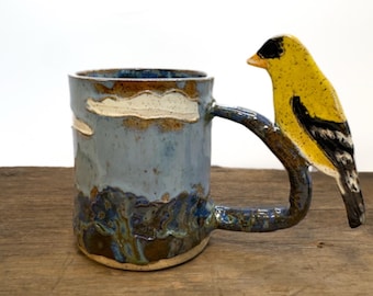 May include: A ceramic mug with a light blue and brown glaze featuring a textured landscape design. The handle is shaped like a tree branch with a yellow, black, and white goldfinch perched on top, extending outward from the side of the mug.
