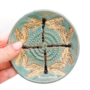 May include: A teal ceramic dish with four dragonfly designs. The dragonflies are embossed and have black bodies and brown wings.
