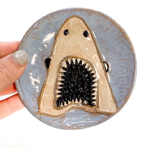 May include: A ceramic plate with a blue glaze and a white shark with a black open mouth. The shark is painted in a cartoon style with black eyes.