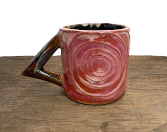 May include: A ceramic mug with a pink exterior featuring a raised spiral pattern and a dark brown, angular, triangular-shaped handle. The interior of the mug is glazed in a dark, glossy black finish, contrasting with the textured pink outer surface.