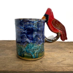 May include: A ceramic mug featuring a glossy glaze with a deep blue upper section and a textured green and brown lower section. The handle is sculpted into the shape of a bright red cardinal bird with a black face mask and an orange beak.