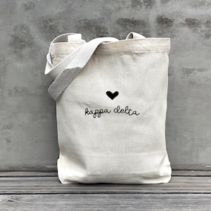 May include: A cream-colored tote bag with a black heart and the words "kappa delta" in a cursive font. The bag has a rectangular shape with a handle and a shoulder strap.