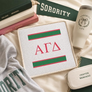 May include: A square needlepoint canvas featuring the Greek letters ΑΓΔ in red, bordered by two horizontal green and red stripes. The canvas is surrounded by a stack of books, a green felt pennant with the text SORORITY, a white tumbler, and a pouch.