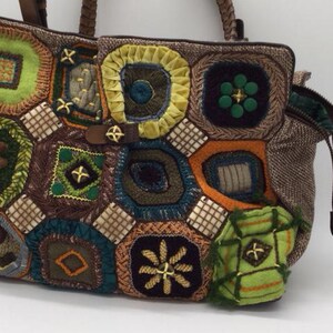 Puede incluir: Un bolso tote de patchwork marr&oacute;n con una variedad de cuadrados de ganchillo de colores. Los cuadrados son en tonos de verde, azul, amarillo, naranja y marr&oacute;n. El bolso tiene cierre de cremallera y asa superior.