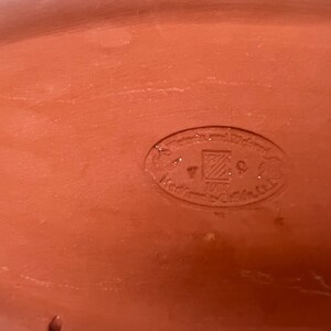 May include: Close-up of a terracotta-colored oval dish with a circular stamp in the center. The stamp reads "Handgefertigt Pottery" in German.