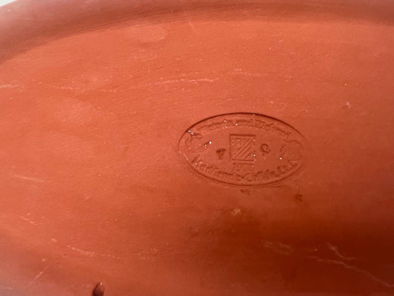 May include: Close-up of a terracotta-colored oval dish with a circular stamp in the center. The stamp reads "Handgefertigt Pottery" in German.