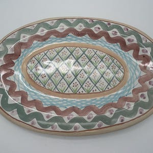 May include: An oval ceramic serving platter with a hand-painted design. The platter features a central oval with a diamond pattern in shades of green, pink, and white. The outer rim is decorated with a wavy pattern in shades of green, brown, and pink.