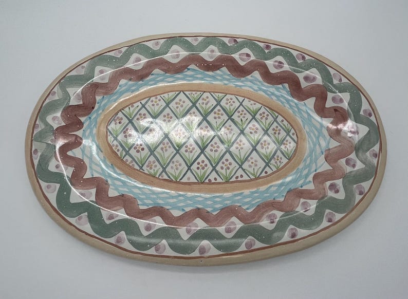 May include: An oval ceramic serving platter with a hand-painted design. The platter features a central oval with a diamond pattern in shades of green, pink, and white. The outer rim is decorated with a wavy pattern in shades of green, brown, and pink.