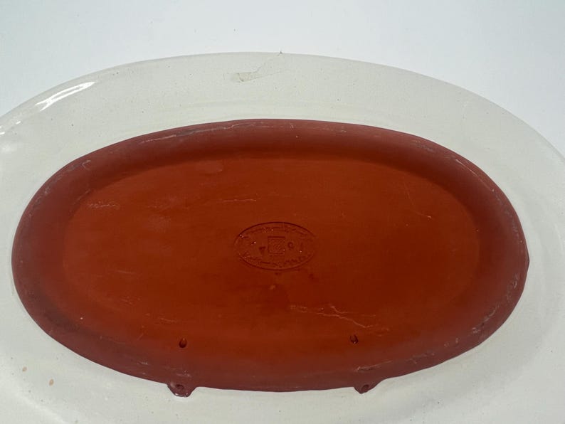 May include: A white oval ceramic serving dish with a red terracotta insert. The insert has a small round stamp with the text "MADE IN ITALY".