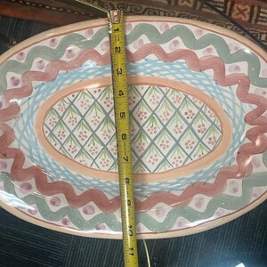 May include: An oval ceramic serving dish with a colorful design. The dish has a light pink background with a blue and green border. The center of the dish has a diamond pattern with pink and green accents.