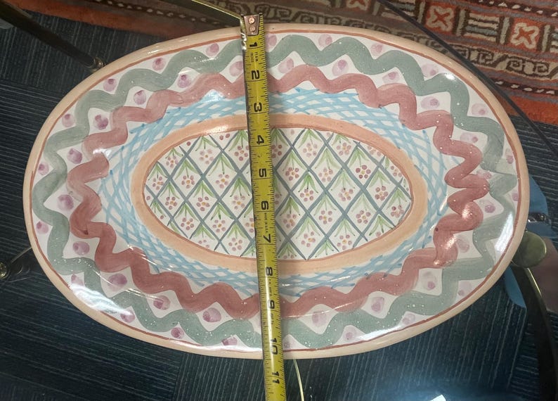 May include: An oval ceramic serving dish with a colorful design. The dish has a light pink background with a blue and green border. The center of the dish has a diamond pattern with pink and green accents.