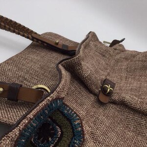 Puede incluir: Un bolso tote tejido marr&oacute;n con correas de cuero y detalles bordados coloridos. El bolso tiene un cierre de solapa con una hebilla de metal.