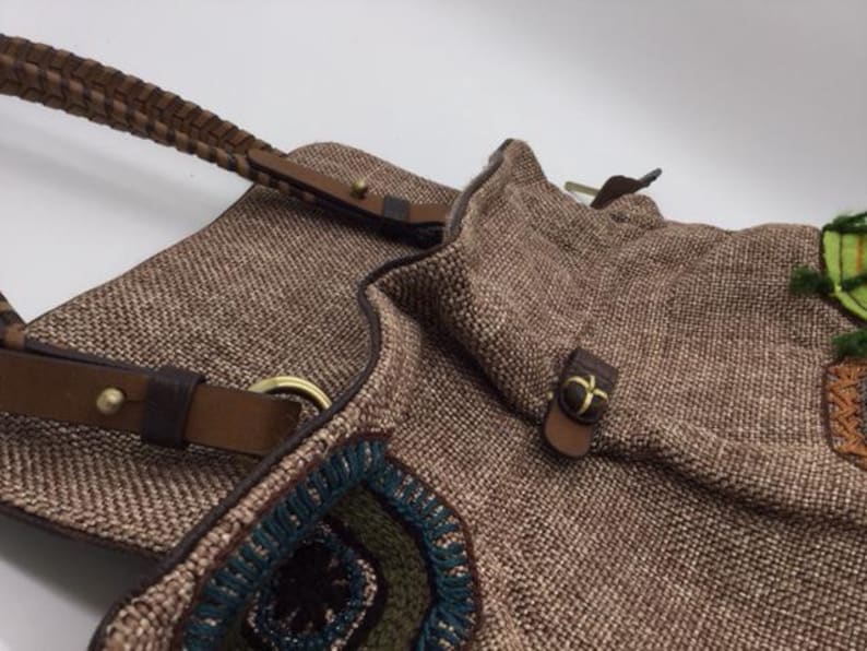 Puede incluir: Un bolso tote tejido marr&oacute;n con correas de cuero y detalles bordados coloridos. El bolso tiene un cierre de solapa con una hebilla de metal.