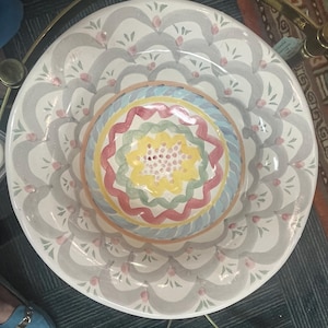 May include: A large, white ceramic serving bowl with a colorful, hand-painted design. The bowl features a central circle with concentric rings of red, green, yellow, and blue. The outer rim of the bowl is decorated with a repeating pattern of gray, white, and pink.