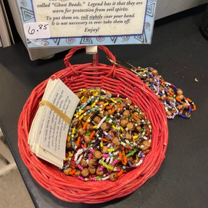 May include: A red woven basket filled with beaded bracelets. The bracelets are made with cedar berries and colorful beads. A sign above the basket reads "Ghost Beads" and describes their protective properties.