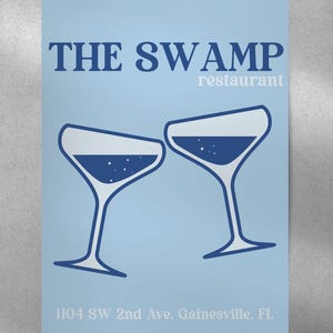 May include: A blue and white poster with the text "THE SWAMP restaurant" and two blue martini glasses clinking together. The address "1104 SW 2nd Ave, Gainesville, FL" is printed at the bottom.