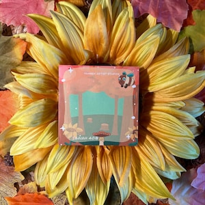 May include: A square card with a nature scene and the text "Isaiah 40:8" is centered on a large yellow and orange sunflower. The card also features a cartoon bee and the text "YAHWEH ARTIST STUDIOS". The background includes colorful fall leaves.