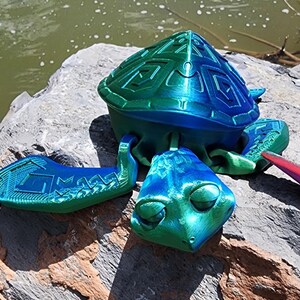 May include: A 3D-printed turtle figurine in shades of blue and green. The turtle has a detailed shell and flippers, with a textured design. The head is visible, and the figurine is resting on a rock.