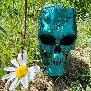 May include: A teal skull-shaped plant pot with visible sutures and cracks, set in grass with a white daisy. The skull has large eye sockets and a detailed jawline, creating a gothic aesthetic. The teal colour gives it a striking appearance.