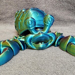 May include: A 3D-printed crab figurine in shades of blue and green. The crab has large claws, a detailed shell, and expressive eyes. The figurine is designed with a gradient color effect, adding depth and visual interest.
