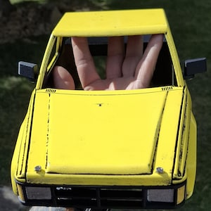 May include: A bright yellow model car body shell featuring a boxy design, black side mirrors, a black front grille, and visible weathering details along the edges. The exterior has a matte finish with metallic silver accents on the front bumper area.