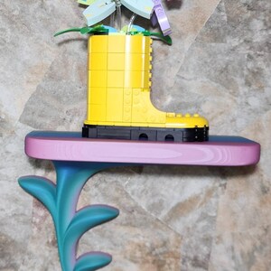 May include: A yellow plastic boot constructed from building blocks serves as a vase for a bouquet of colorful plastic flowers. The boot sits on a horizontal shelf with a pink top and a teal, leaf-shaped support bracket extending downward.