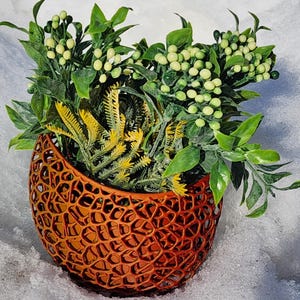 Peut inclure: Un pot orange à tissage ouvert rempli de verdure artificielle. Le pot a un design texturé unique. La verdure comprend diverses nuances de vert et de jaune, avec de petits accents blancs ronds. Le pot est posé sur un fond enneigé.