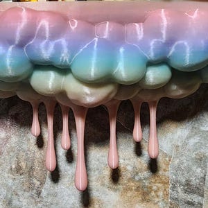 May include: A wall-mounted shelf shaped like a stylized cloud with a gradient finish transitioning from soft pink to blue and purple. The bottom edge of the cloud features several smooth, light pink drips extending downward in a vertical pattern.
