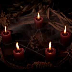 May include: Five red candles with lit flames arranged in a circle, surrounded by a rustic arrangement of rope, dried leaves, and a spiderweb. The candles are the focal point, casting a warm glow against the dark background, creating a mystical ambiance.