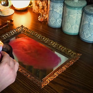K&ouml;nnte beinhalten: Ein goldgerahmter Spiegel reflektiert ein wirbelndes rot-rosa Design. Drei dekorative Gl&auml;ser mit blau-wei&szlig;en Mustern stehen auf einer Holzoberfl&auml;che. Eine kleine Flamme brennt in einer schwarzen Schale.