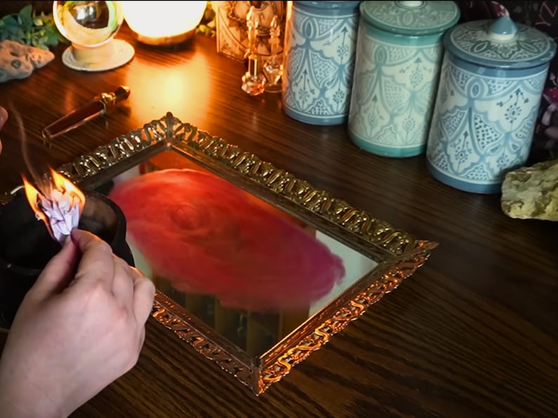K&ouml;nnte beinhalten: Ein goldgerahmter Spiegel reflektiert ein wirbelndes rot-rosa Design. Drei dekorative Gl&auml;ser mit blau-wei&szlig;en Mustern stehen auf einer Holzoberfl&auml;che. Eine kleine Flamme brennt in einer schwarzen Schale.