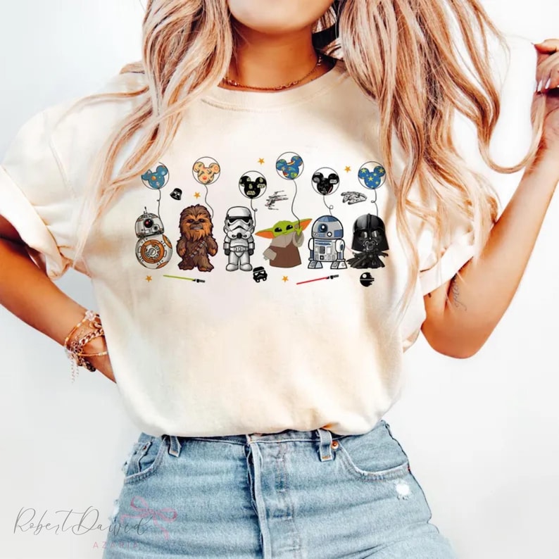 K&ouml;nnte beinhalten: Cremefarbenes T-Shirt mit Star Wars-Charakteren als Ballons. Das Design umfasst BB-8, Chewbacca, einen Sturmtruppler, Grogu, R2-D2 und Darth Vader. Die Ballons haben Mickey Mouse Ohren. Das T-Shirt wird von einer Person getragen.
