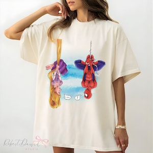 K&ouml;nnte beinhalten: Cremefarbenes T-Shirt mit einer Cartoon-Illustration von Rapunzel und Spider-Man, die kopf&uuml;ber h&auml;ngen. Rapunzel h&auml;lt ihr langes blondes Haar, und Spider-Man h&auml;ngt an einer Kette. Das Shirt hat den Text "O HAI!" und ein Fragezeichen.