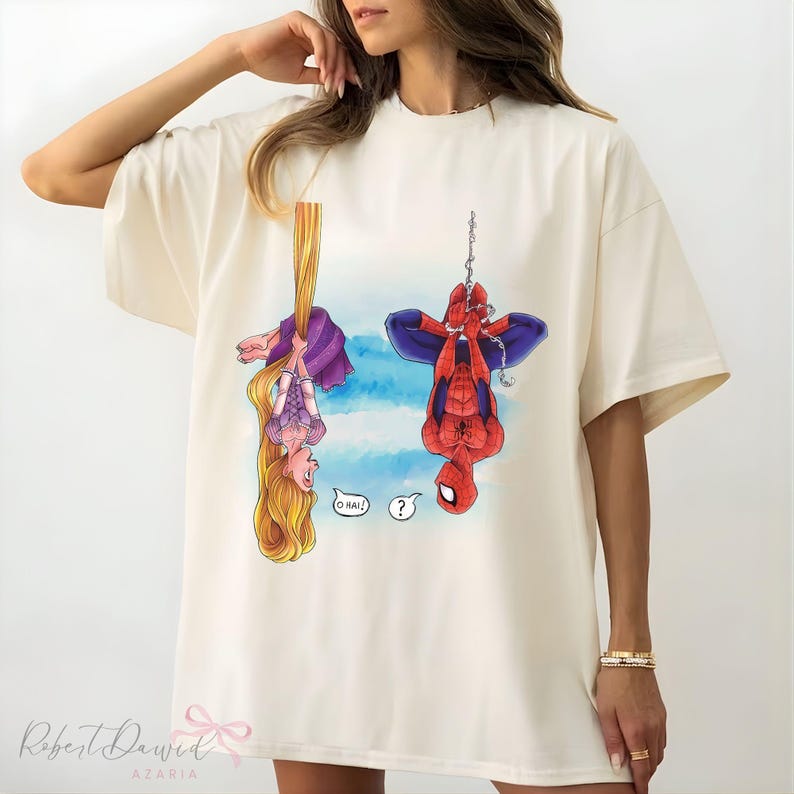 K&ouml;nnte beinhalten: Cremefarbenes T-Shirt mit einer Cartoon-Illustration von Rapunzel und Spider-Man, die kopf&uuml;ber h&auml;ngen. Rapunzel h&auml;lt ihr langes blondes Haar, und Spider-Man h&auml;ngt an einer Kette. Das Shirt hat den Text "O HAI!" und ein Fragezeichen.