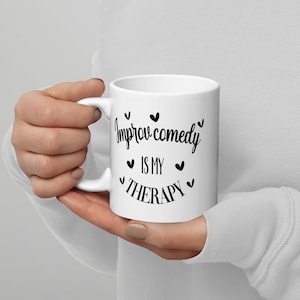 May include: White ceramic mug with black text that reads "Improv comedy is my therapy" with small hearts around the text.
